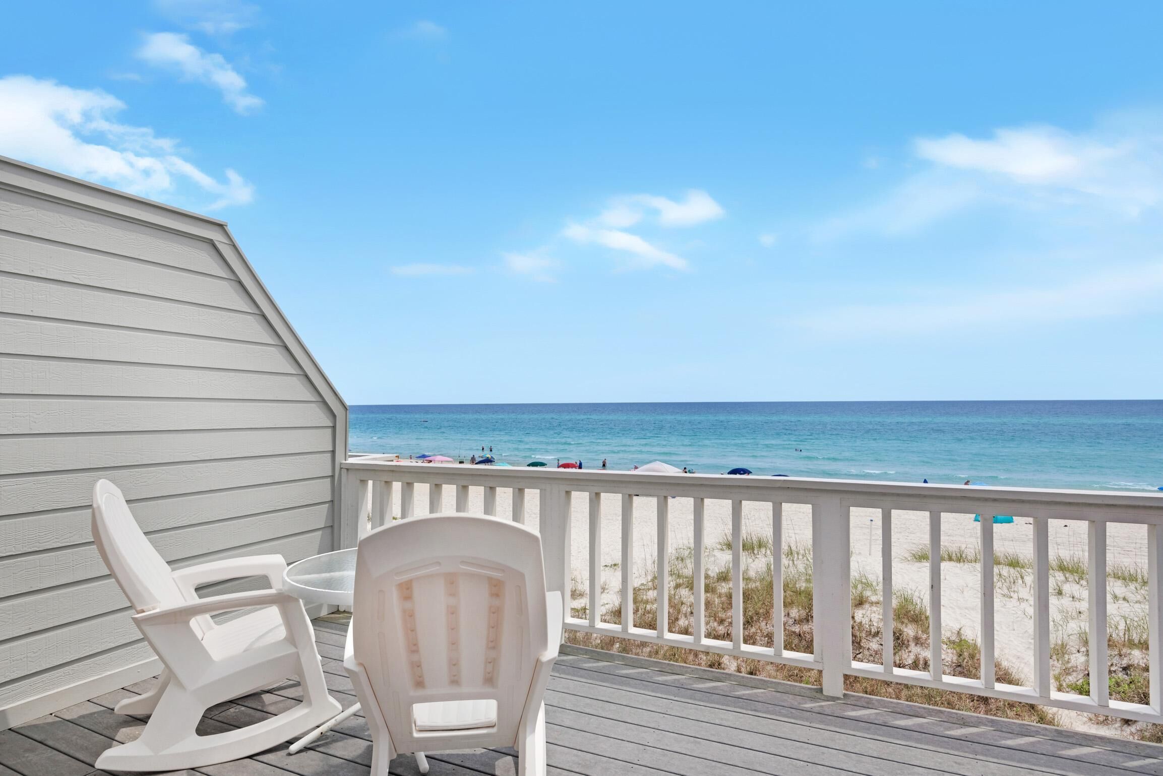 A deck with white chairs and a table overlooking a beach with people and umbrellas.