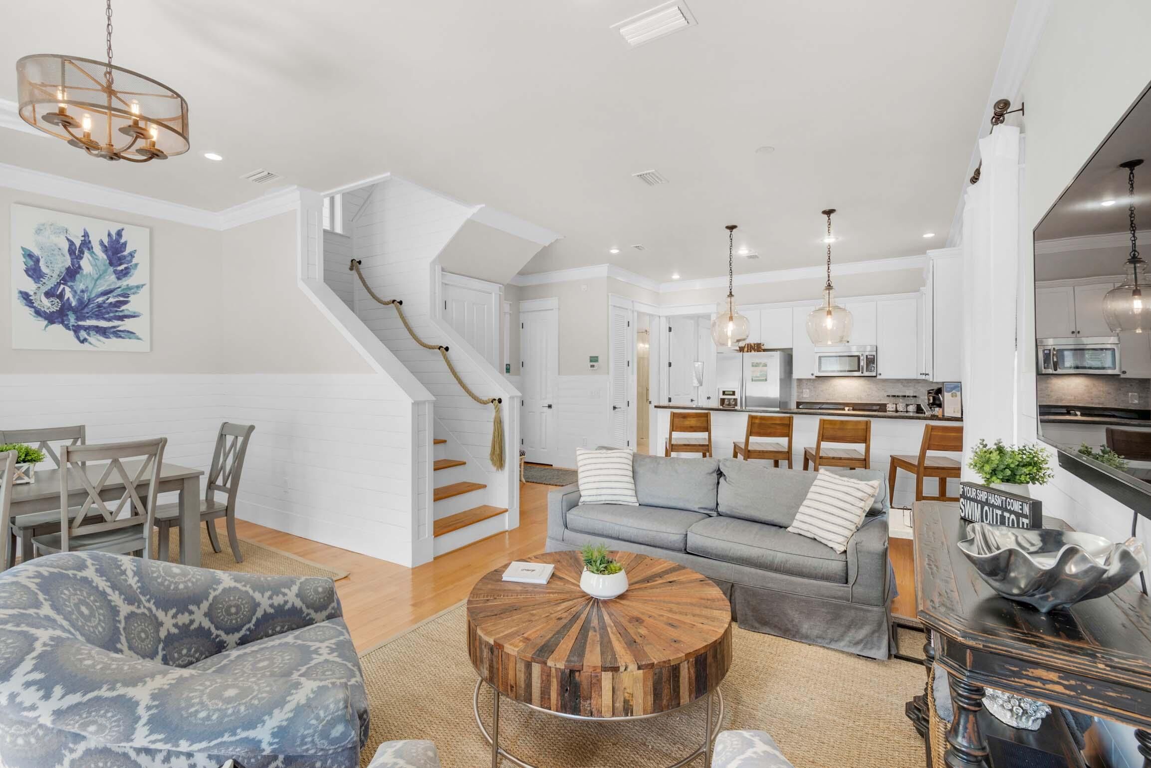 A spacious living area with a gray sofa, a round wooden coffee table, and a dining table with chairs. A staircase leads to an upper level.