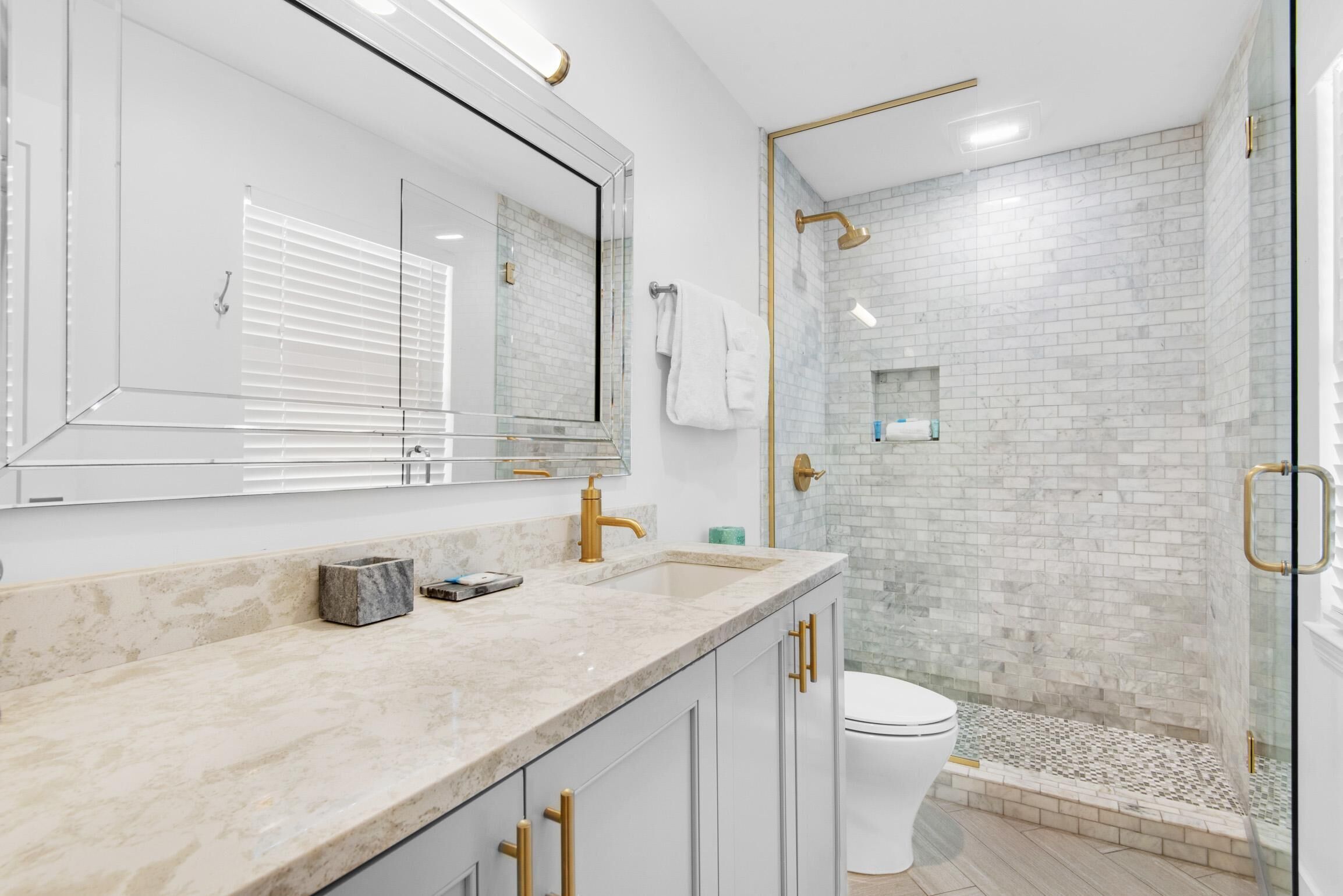 A modern bathroom with a large mirror, a marble countertop, a white toilet, and a glass-enclosed shower area.