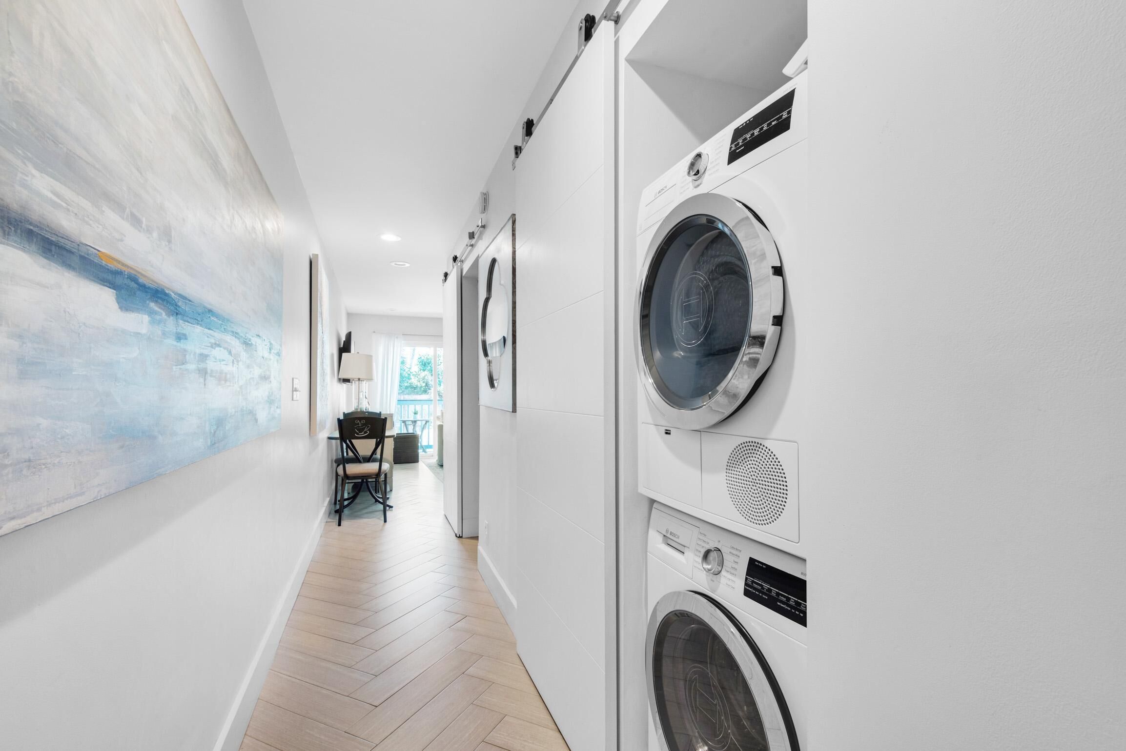 A laundry room with a large wall-mounted washing machine and dryer, a wooden floor, and a view into a dining area with chairs and a table.