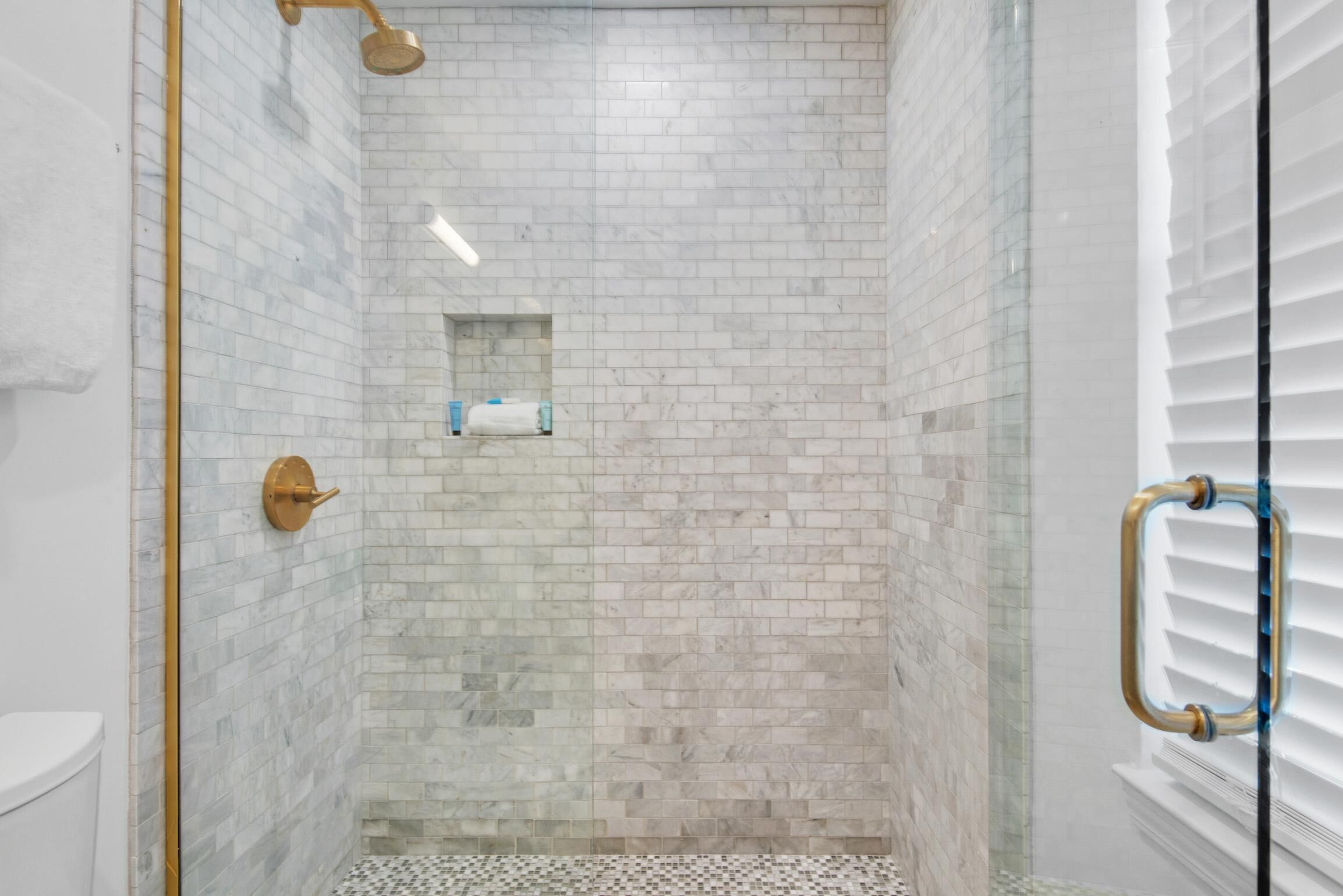 A modern shower with a glass door, white marble tiles, and a gold faucet.