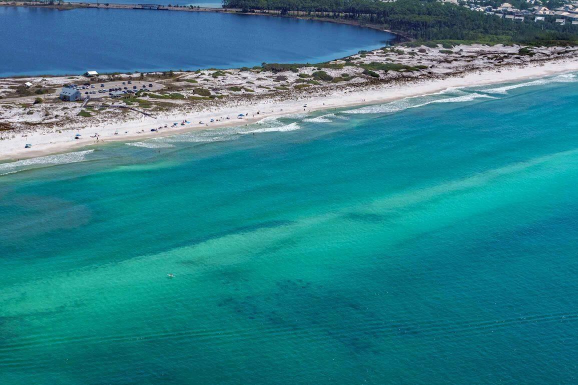 Vista aérea de una playa con aguas turquesas cristalinas, una orilla arenosa y algunos edificios al fondo.