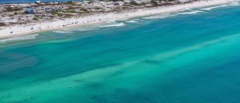 Vista aérea de una playa con aguas turquesas cristalinas, una orilla arenosa y algunos edificios al fondo.