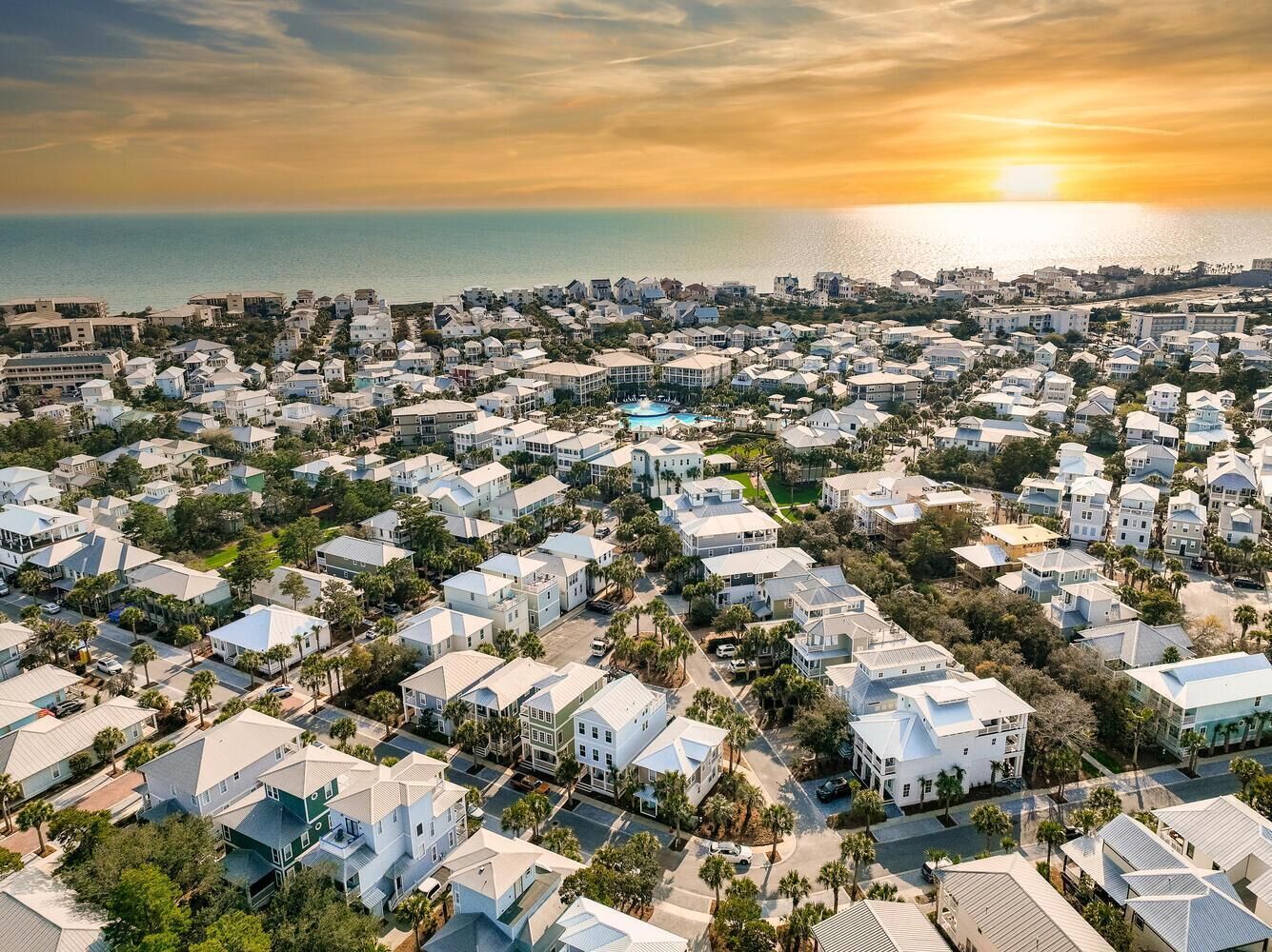 An aerial view of a coastal residential area with uniform housing, a swimming pool, and a sunset over the ocean.