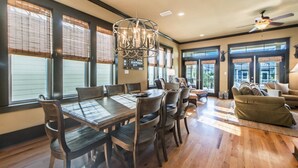 A dining area with a wooden table and chairs, a chandelier, and large windows with blinds. - Comfort and Joy (Panama City Beach)