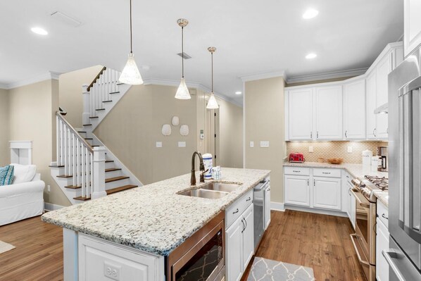 A modern kitchen with white cabinetry, granite countertops, and a central island with a sink. There is a staircase leading to another level, and the kitchen is well-lit with pendant lights.