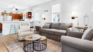 Una sala de estar moderna con un sofá, sillones y una mesa de centro. Hay una zona de cocina con armarios de madera y un refrigerador.