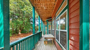 A wooden deck with a bench, surrounded by trees and a building with green and red siding. - Playhouse II (Santa Rosa Beach)