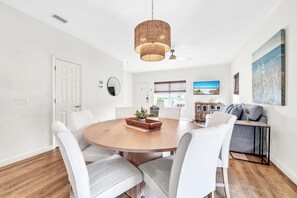 A dining area with a round wooden table, white upholstered chairs, and a woven pendant light. - Palm Grove at Sago Sands Unit A (Santa Rosa Beach)