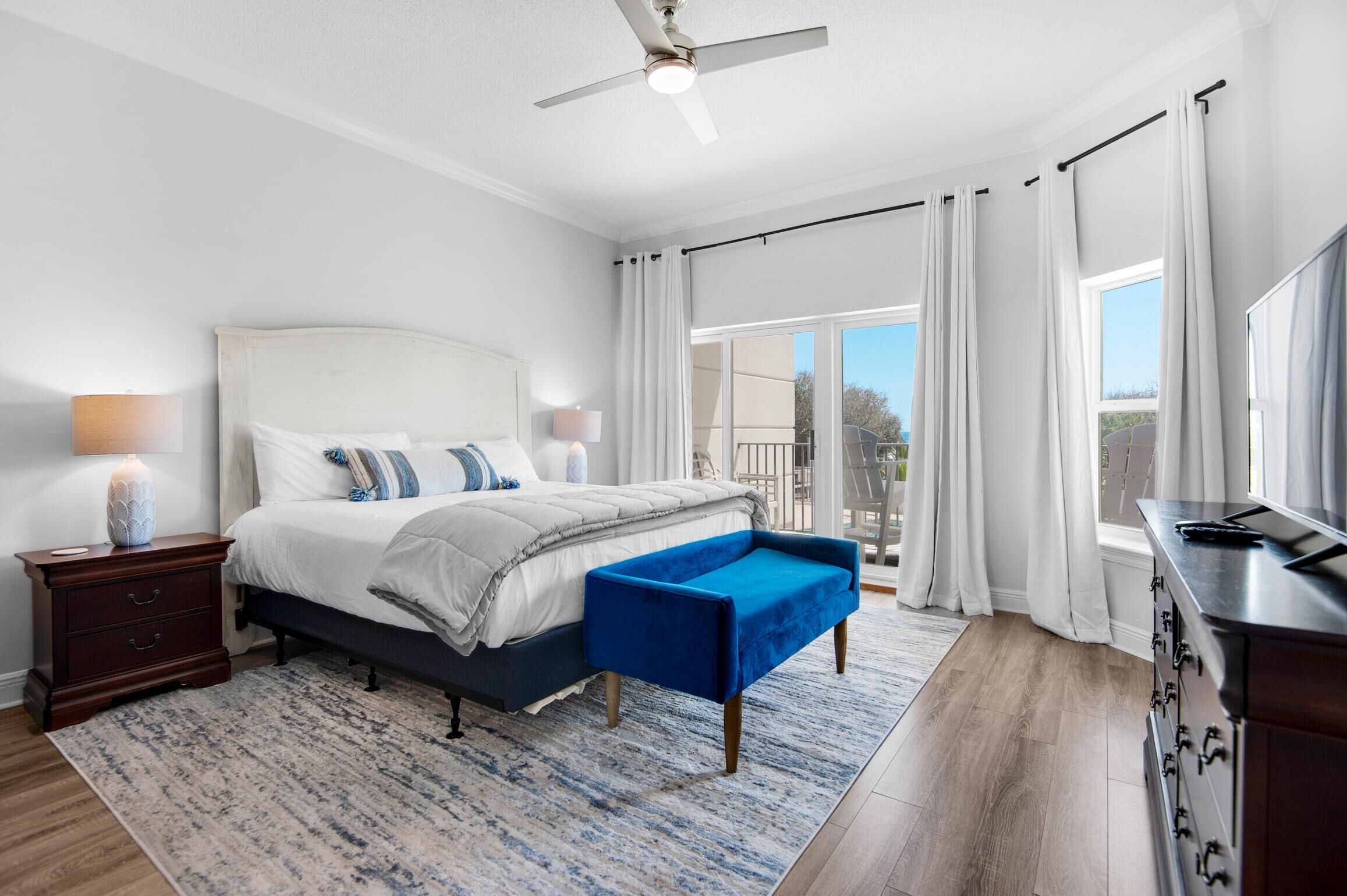 Un dormitorio con cama, mesitas de noche, un banco azul, ventilador de techo y vistas al exterior.