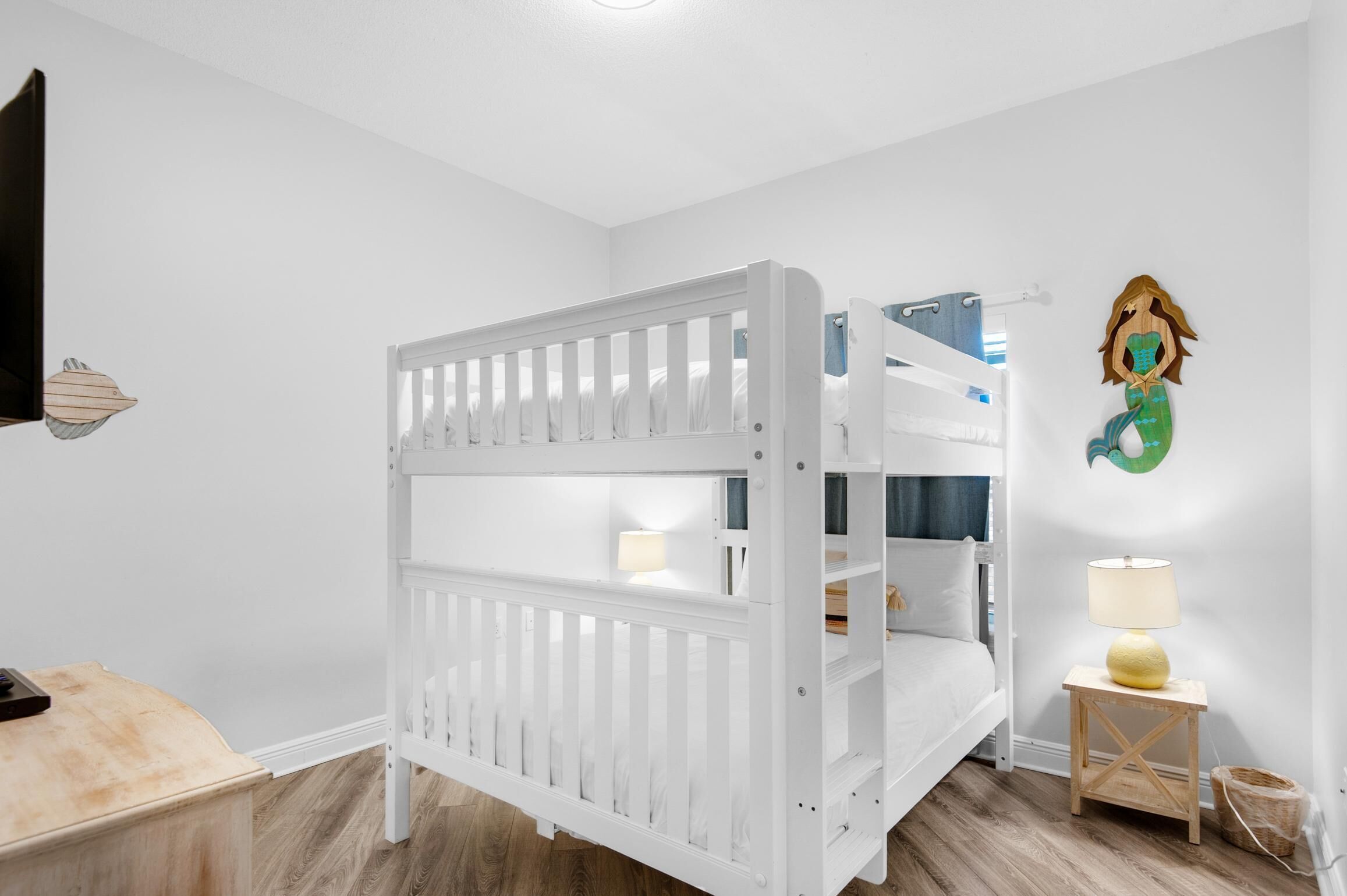 A white bunk bed in a room with a wooden floor, a small bedside table with a lamp, and a wall decoration.