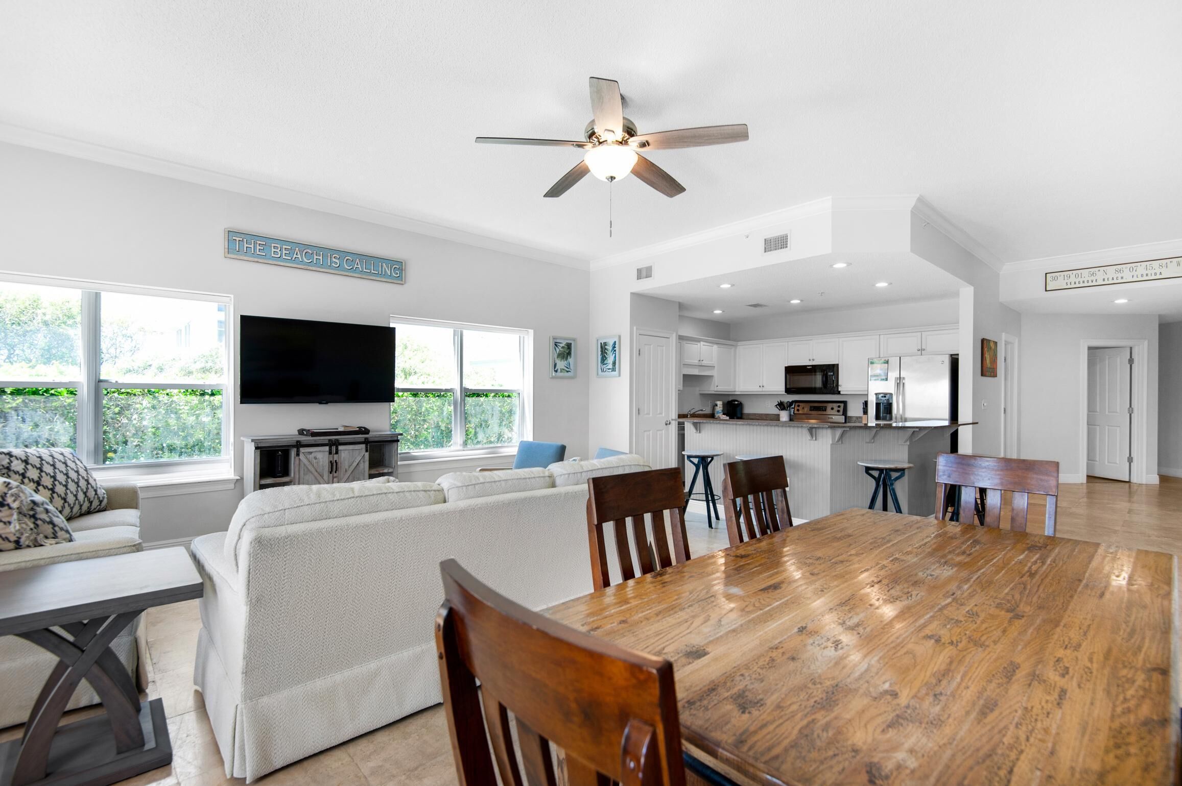 A spacious living area with a large wooden dining table, white sofas, a flat-screen TV, and a ceiling fan.