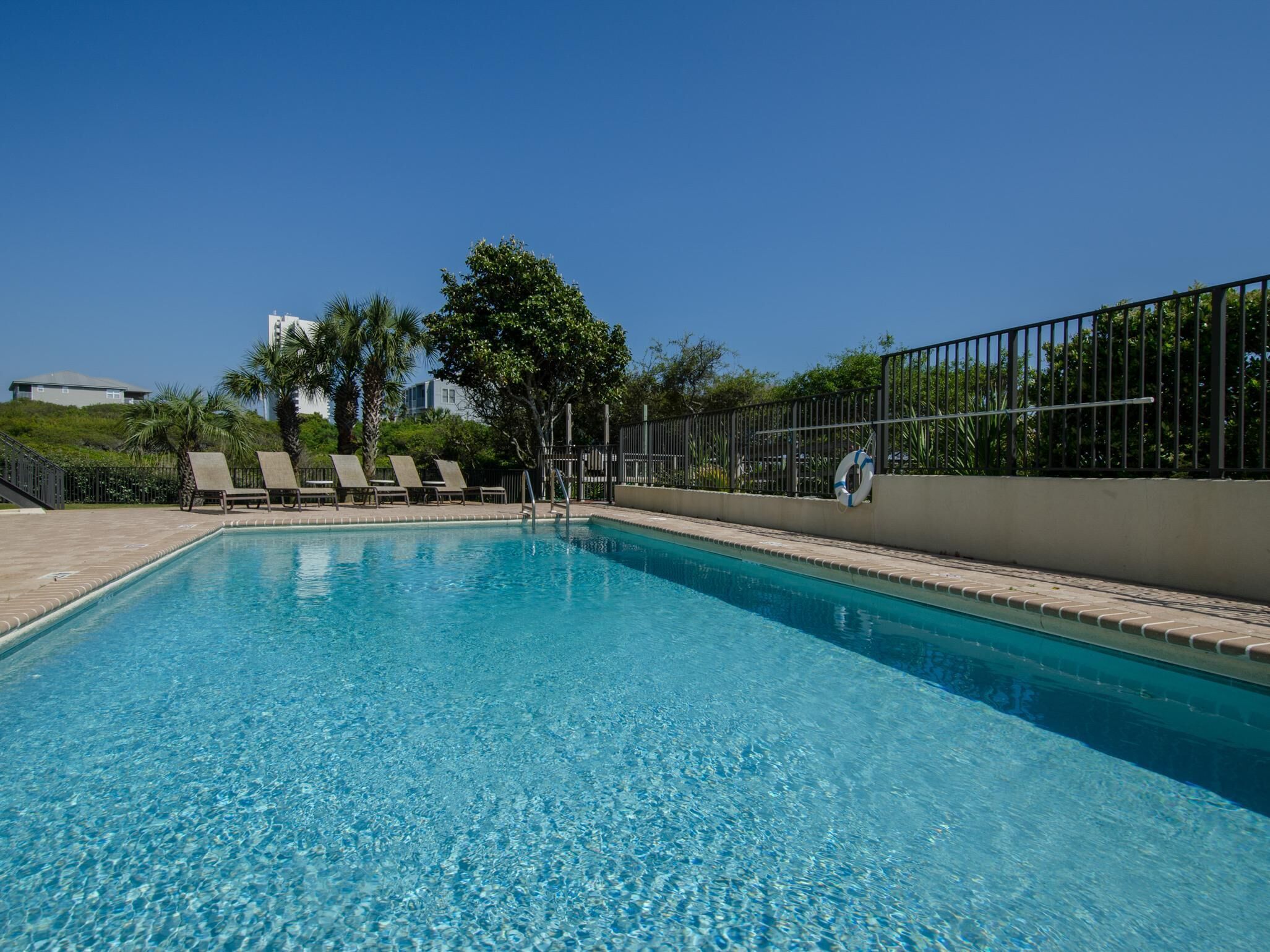 A rectangular swimming pool with clear blue water, surrounded by a low brick wall and a metal fence. There are several lounge chairs aligned along the wall, and a lifebuoy is attached to the fence.