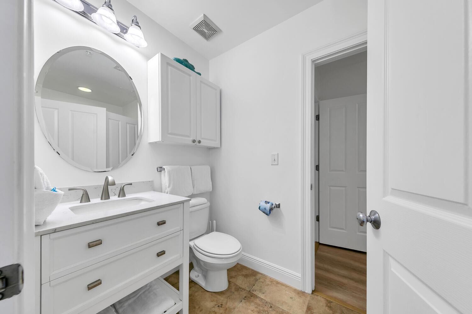 A bathroom with a white vanity, a round mirror, a toilet, and a door leading to another room.