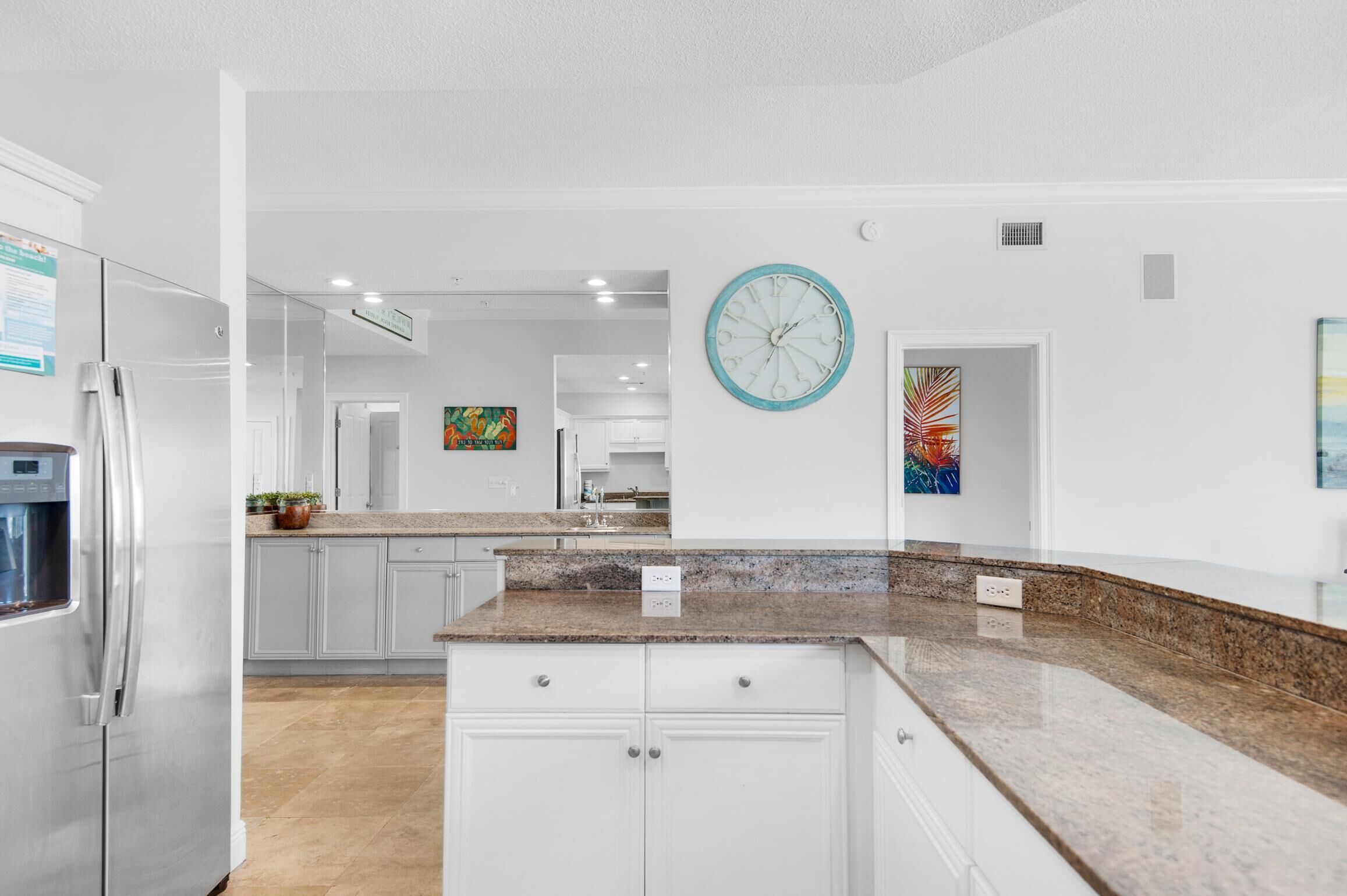 A modern kitchen with white cabinets, granite countertops, a stainless steel refrigerator, and a large wall clock.