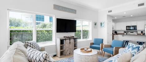 A spacious living room with a large window, a flat-screen TV, and a kitchen area visible through an open door.