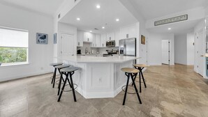 A modern kitchen with white cabinetry, a central island with stools, and a view of the outdoors. - Legacy 104 (Santa Rosa Beach)