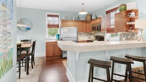 A kitchen with wooden cabinets, a central island, bar stools, and a dining area with chairs. - Four Fish Bungalow 155 (Santa Rosa Beach)