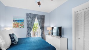 A bedroom with a bed, a TV, a ceiling fan, a window with patterned curtains, and a door. - Four Fish Bungalow 155 (Santa Rosa Beach)