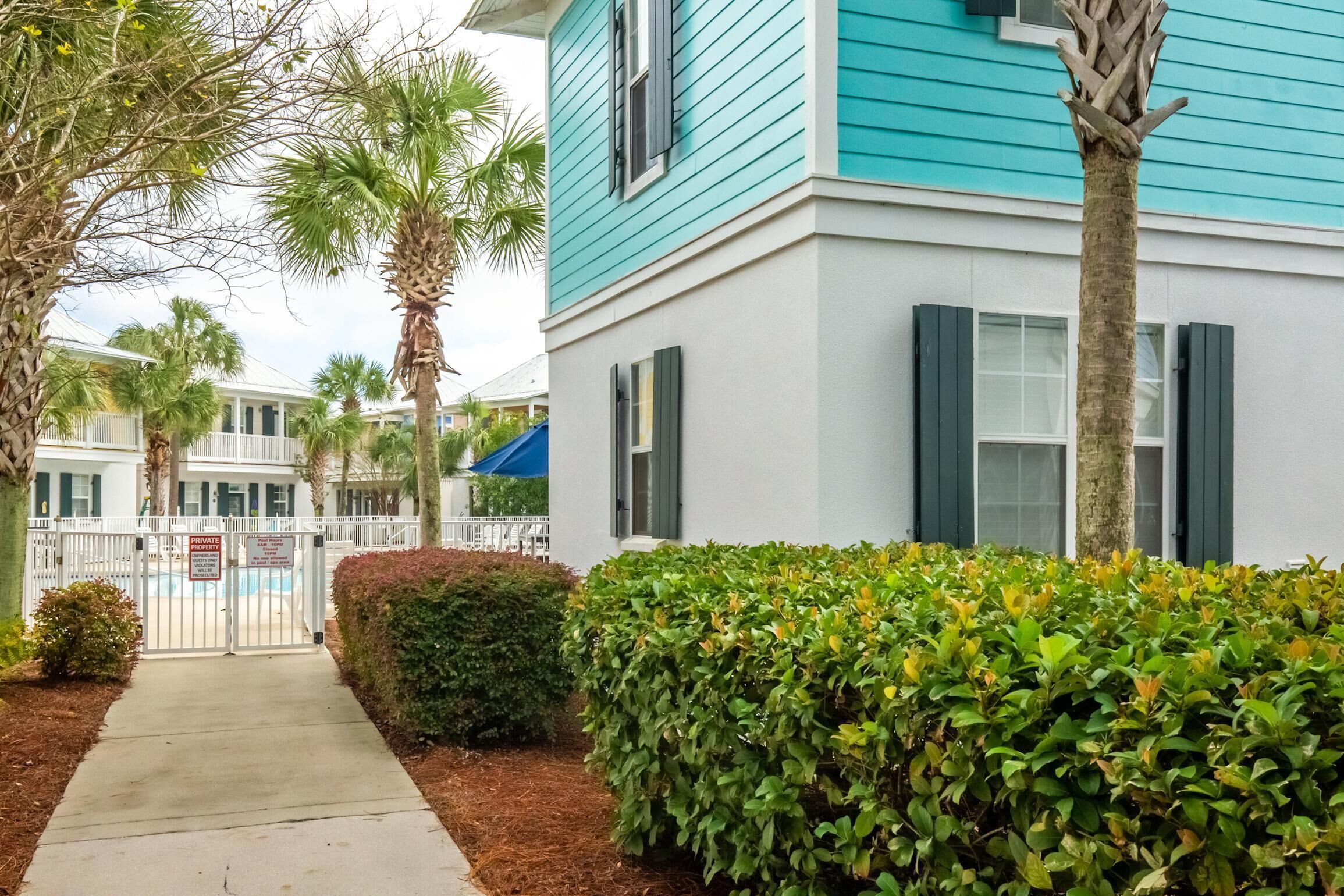 A residential building with a white exterior and teal siding, surrounded by a white fence and palm trees.