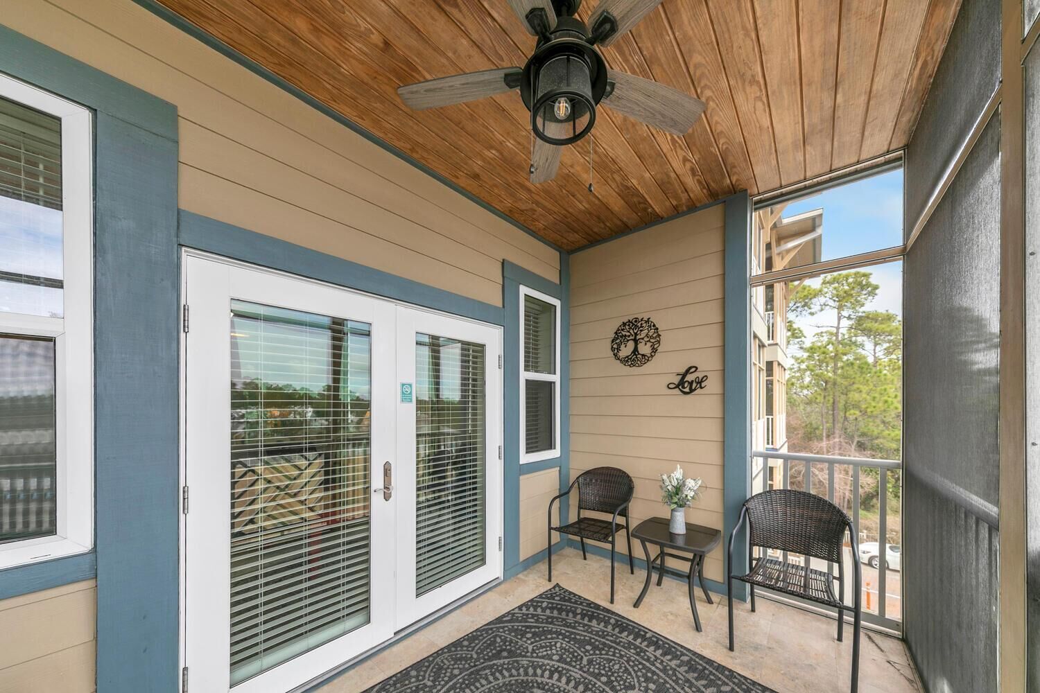 A covered porch with a ceiling fan, two doors with blinds, a small table with chairs, and a decorative wall piece.