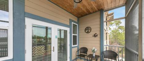 A covered porch with a ceiling fan, two doors with blinds, a small table with chairs, and a decorative wall piece.