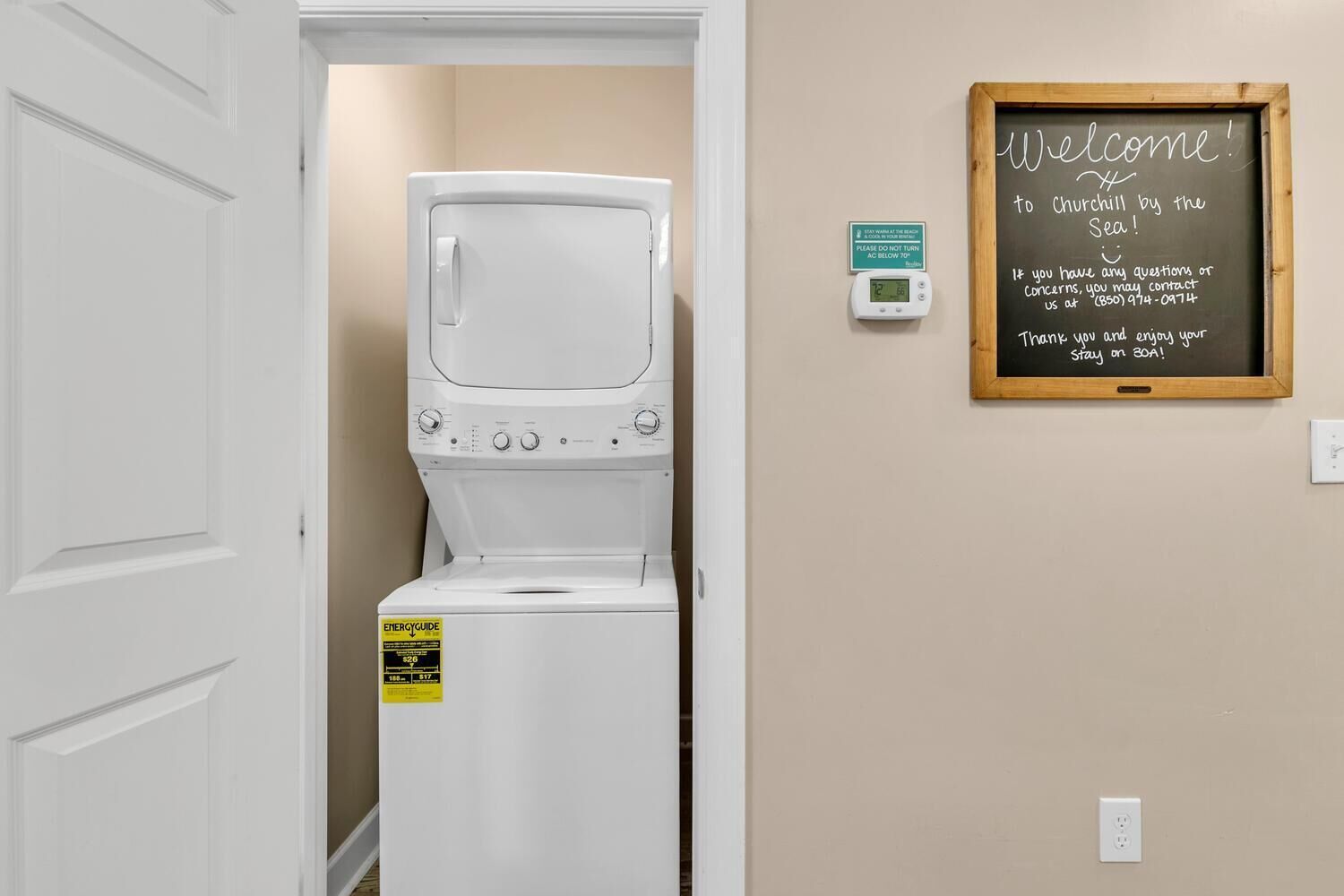 A laundry room with a front-loading washer and dryer, a thermostat, and a welcome sign on the wall.