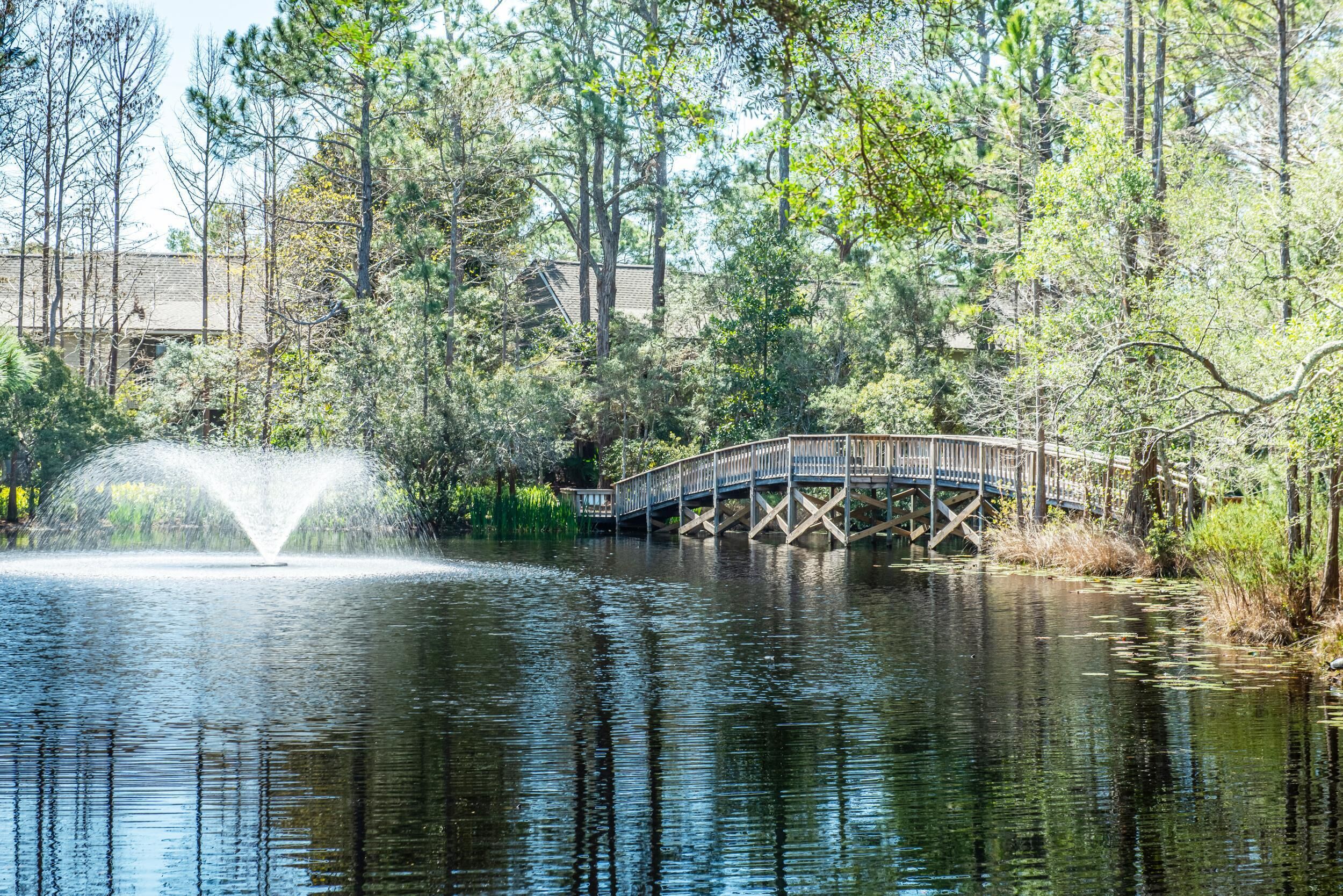 A wooden bridge over a calm body of water with a fountain in the center, surrounded by trees and a building in the background.