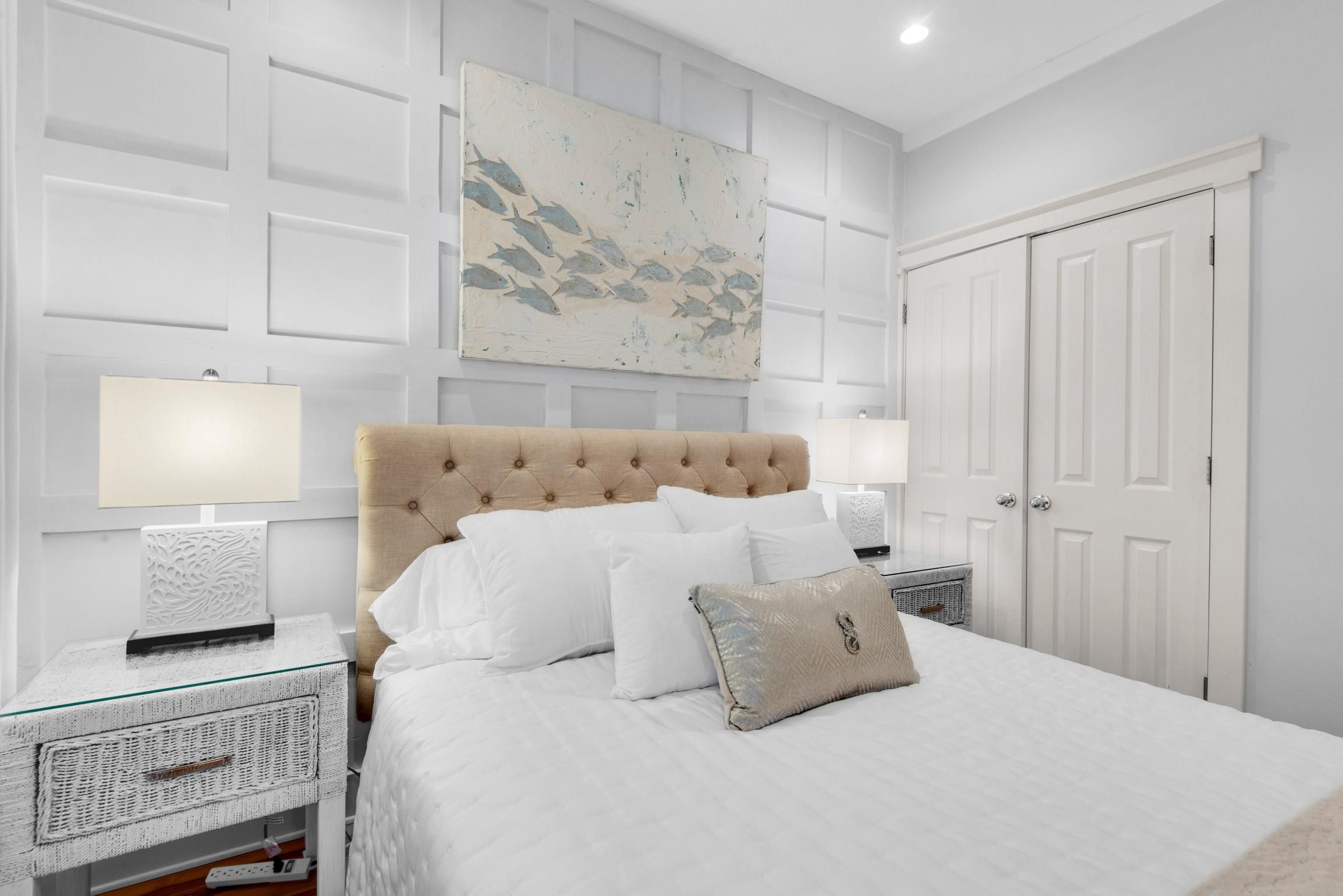 A bedroom with a tufted headboard, white bedding, a glass bedside table, a wall-mounted artwork, and a white door.