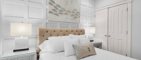 A bedroom with a tufted headboard, white bedding, a glass bedside table, a wall-mounted artwork, and a white door.