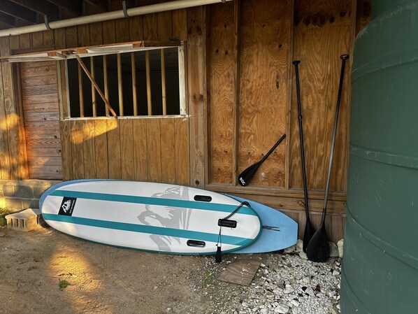 Een surfplank die tegen een houten muur rust, met een raam en een peddel die ook tegen de muur staat.