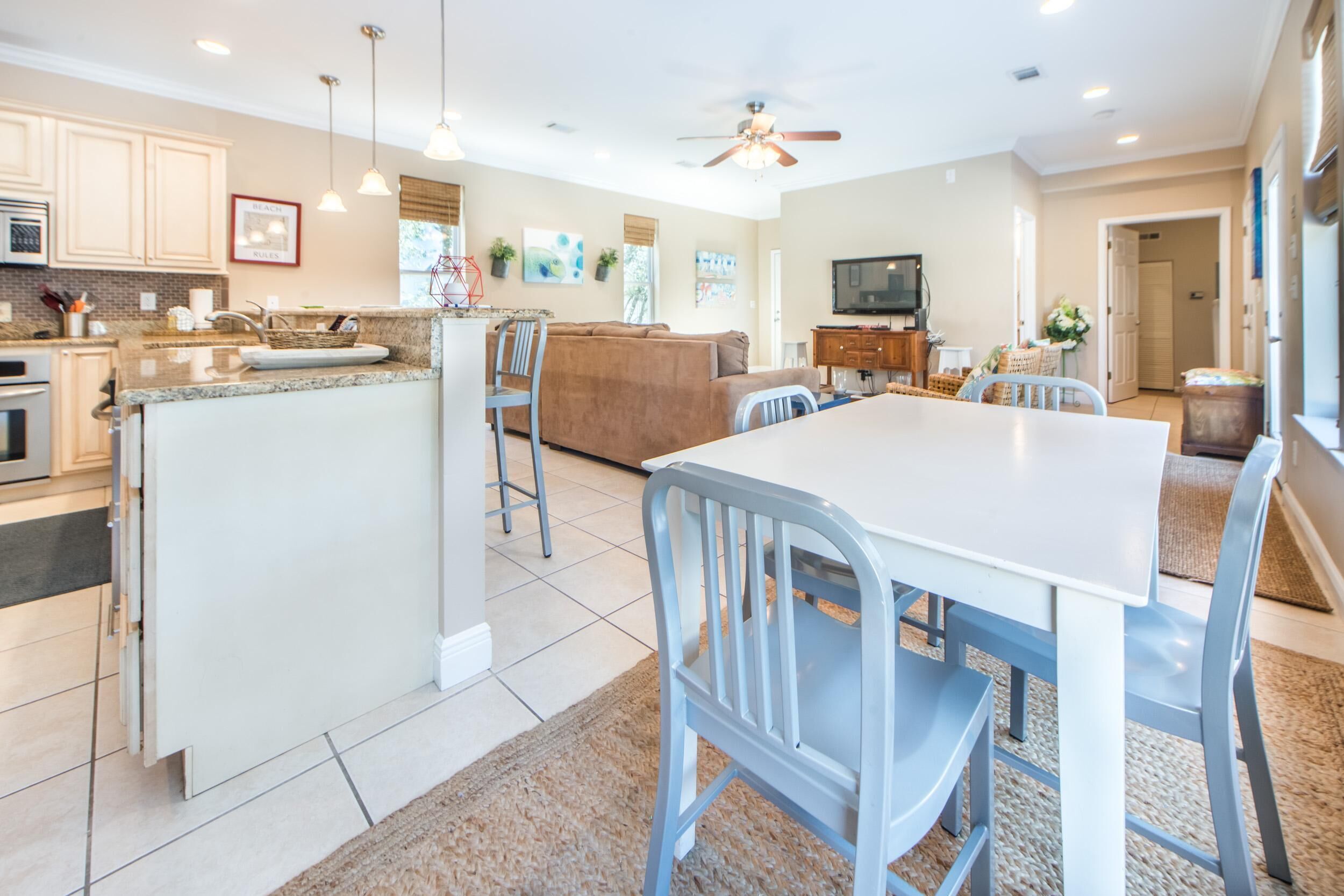 A modern kitchen with granite countertops, a dining area with a white table and blue chairs, and a living room with a brown sofa and a television.