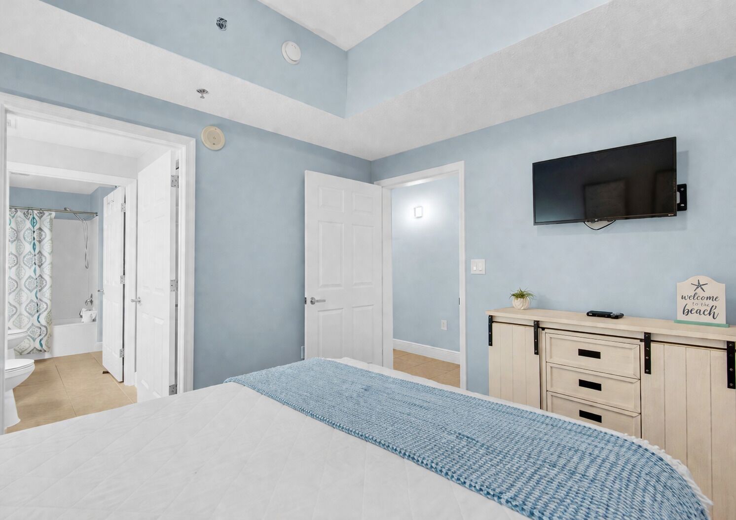 A bedroom with a bed, a TV mounted on the wall, a bedside table with a sign, and a door leading to a bathroom.