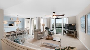A spacious living room with a large sofa, a coffee table, and a television. There is a dining area with a table and chairs, and a ceiling fan. - Enclave 304b (Destin)