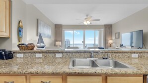 A modern kitchen with granite countertops, wooden cabinets, a double sink, and a large TV. - Ariel Dunes I 1505 (Miramar Beach)