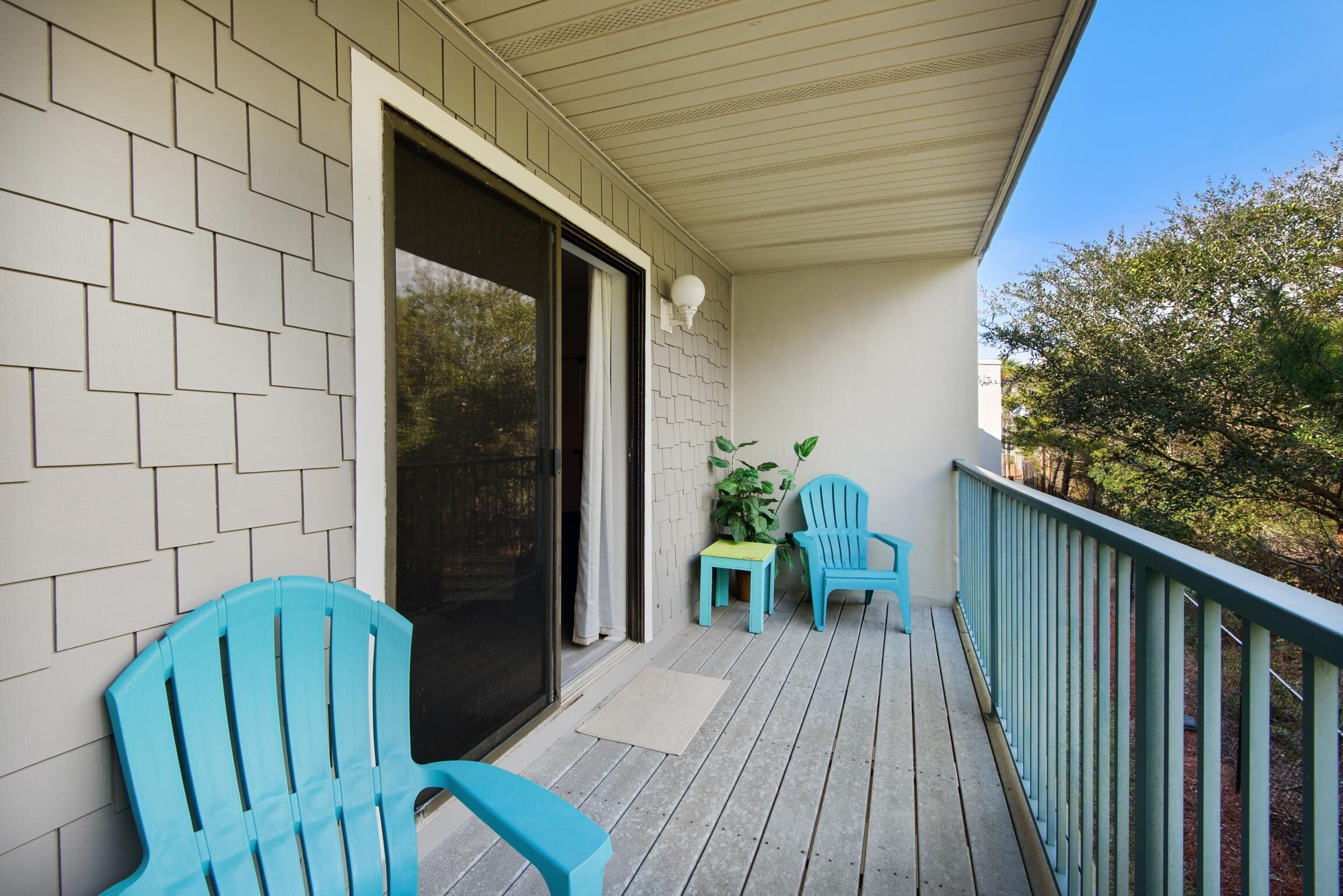 A covered porch with a sliding glass door, two blue Adirondack chairs, a small table, and a potted plant.