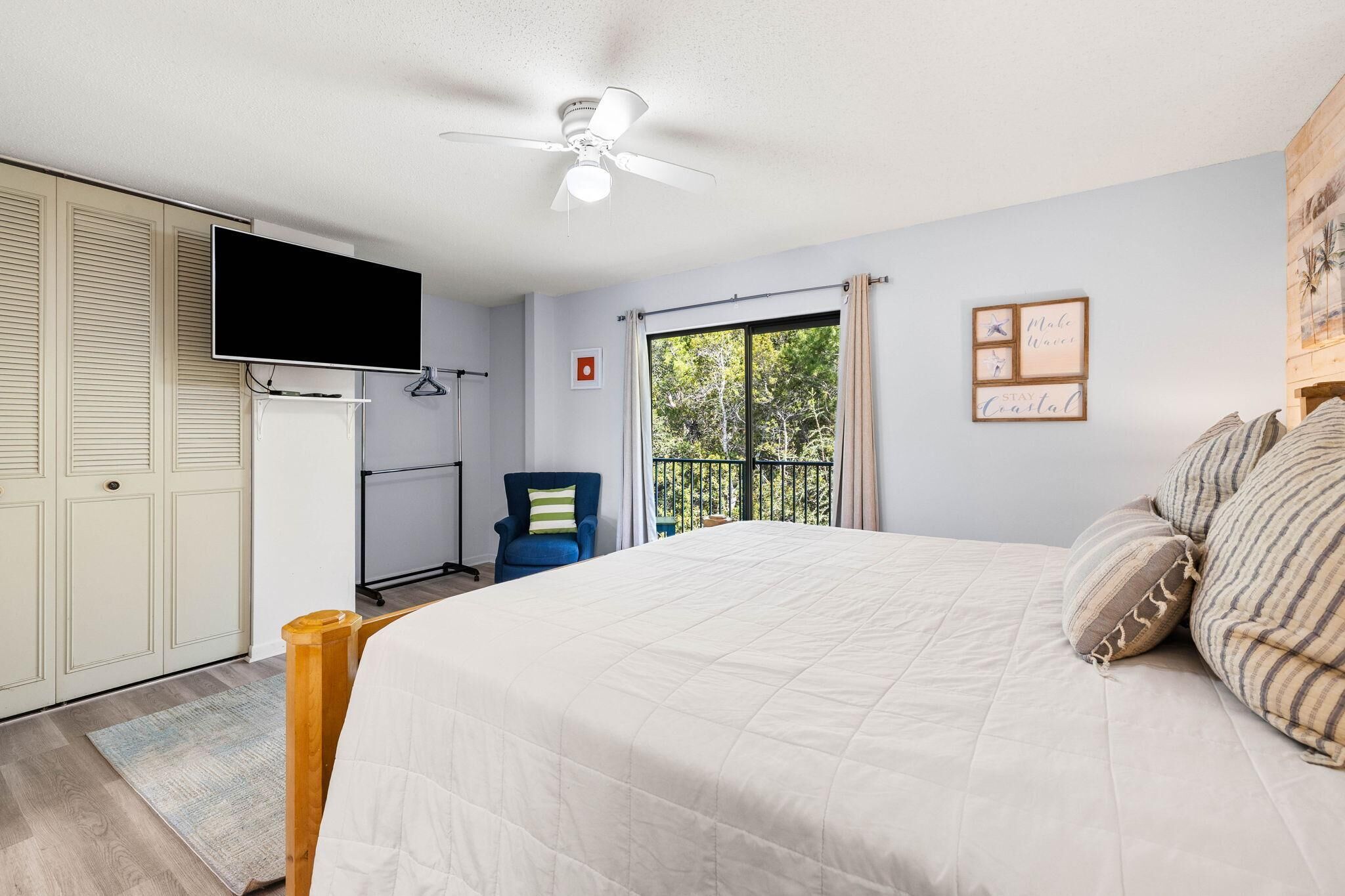 Un dormitorio con cama, ventilador de techo, televisión, silla, ventana con cortinas y balcón.