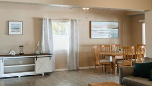 A living room with a wooden dining table, chairs, a sofa, and a kitchen area with a white cabinet and a window with curtains. - Cozy Home 20 min to Beach! Private Backyard. Pet Friendly (Fort Myers)