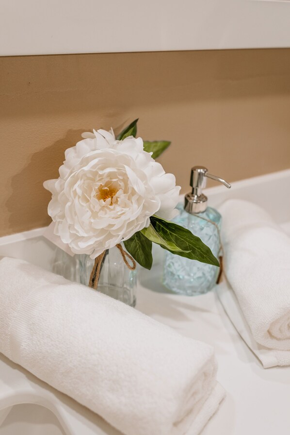A bathroom with a white peony in a glass vase, a soap dispenser, and neatly folded white towels.