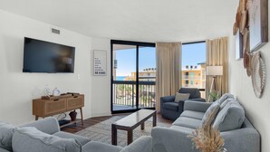 A modern living room with a flat-screen TV, grey sofas, a wooden coffee table, and a large sliding glass door leading to a balcony with a view of buildings and the sea. - Sundestin 416 (Destin)