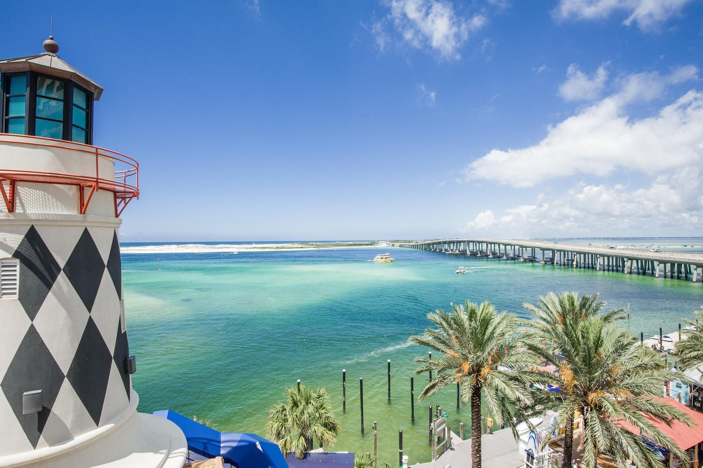 Ein Leuchtturm mit diamantmusterfassadier, eine Pier, die sich in türkisfarbenes Wasser erstreckt, und Palmen im Vordergrund.