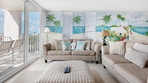 A living room with a beige sofa, a matching ottoman, and a large window overlooking a balcony. - Dolphin Point #406c (Destin)