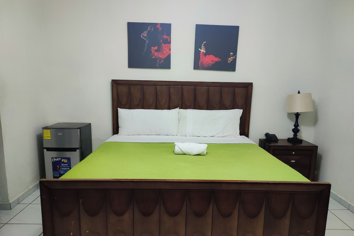 Habitación de hotel con cama de madera, colcha verde, dos almohadas blancas, una mesita de noche con lámpara y un refrigerador.