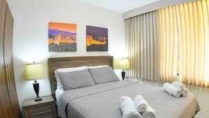 Habitación de hotel con una cama grande, dos mesitas de noche con lámparas, un aparador, un ventanal con cortinas y dos cuadros decorativos.