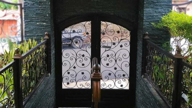 Una puerta negra con un enrejado de metal decorativo, rodeada por una barandilla negra y follaje.
