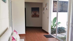 Un pasillo con suelo de madera, un sofá blanco con cojines de colores, una lámpara en el techo y una puerta que da a un balcón con plantas.
