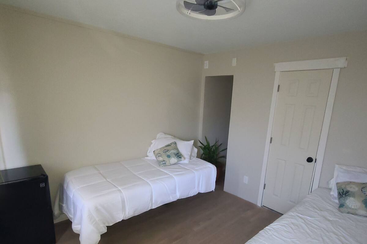 A bedroom with two beds, a ceiling fan, a door, and a small black appliance.