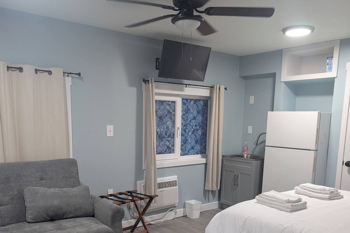 A bedroom with a bed, a grey sofa, a ceiling fan, a window with curtains, a small kitchenette, and a white refrigerator.