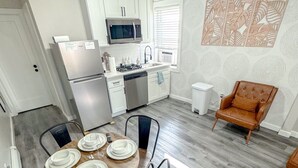 Een moderne keuken met witte kastjes, een roestvrijstalen koelkast en een vaatwasser. Er is een eethoek met een ronde houten tafel voor vier personen, omringd door zwarte stoelen. Een bruine leren fauteuil is op de achtergrond zichtbaar.
