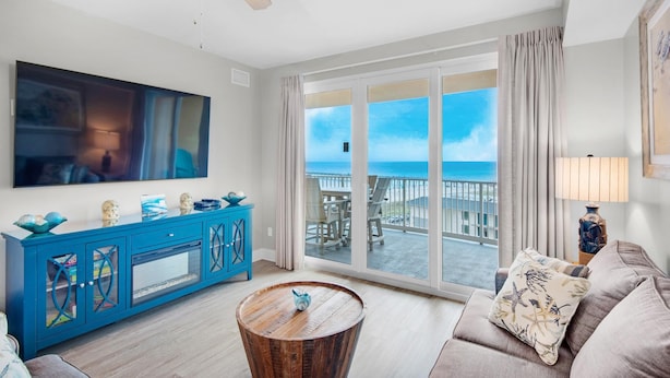 Een woonkamer met een grote televisie, een blauwe kast, een ronde houten salontafel en uitzicht op de oceaan door grote ramen.